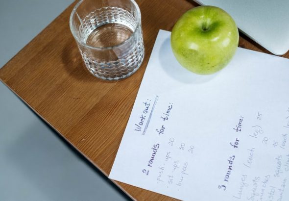 A green apple, glass of water, and workout plan on a wooden desk symbolize healthy living.